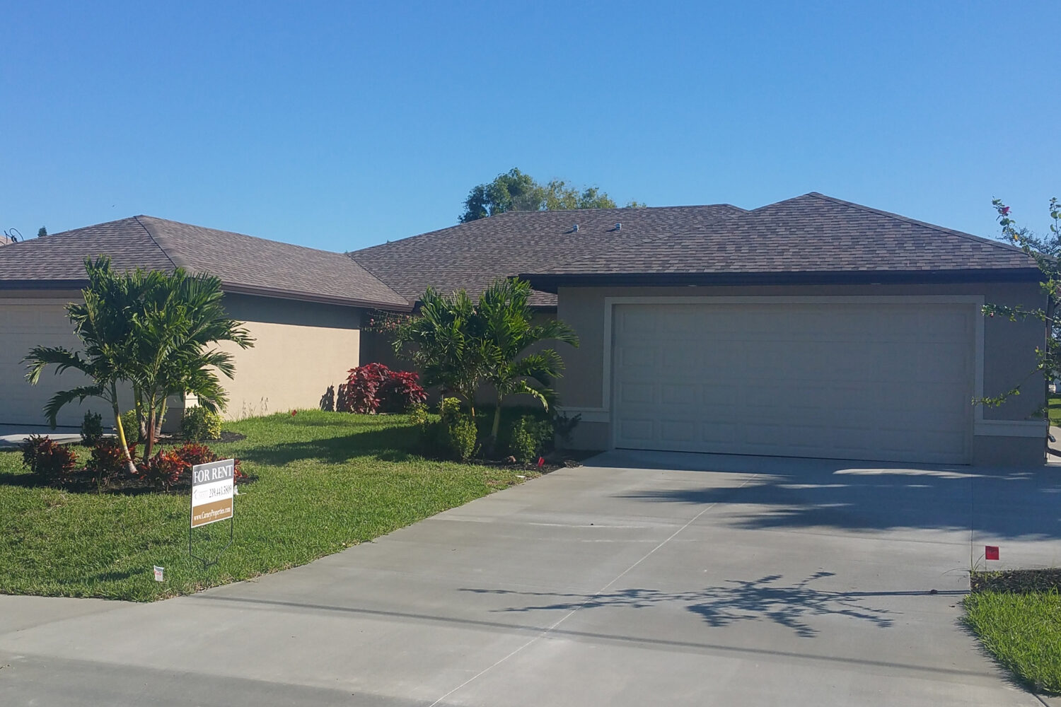 Single-story house with a double garage, palm trees, and a small lawn under a clear blue sky. A real estate sign in the front yard highlights this inviting option among homes for rent in Naples.