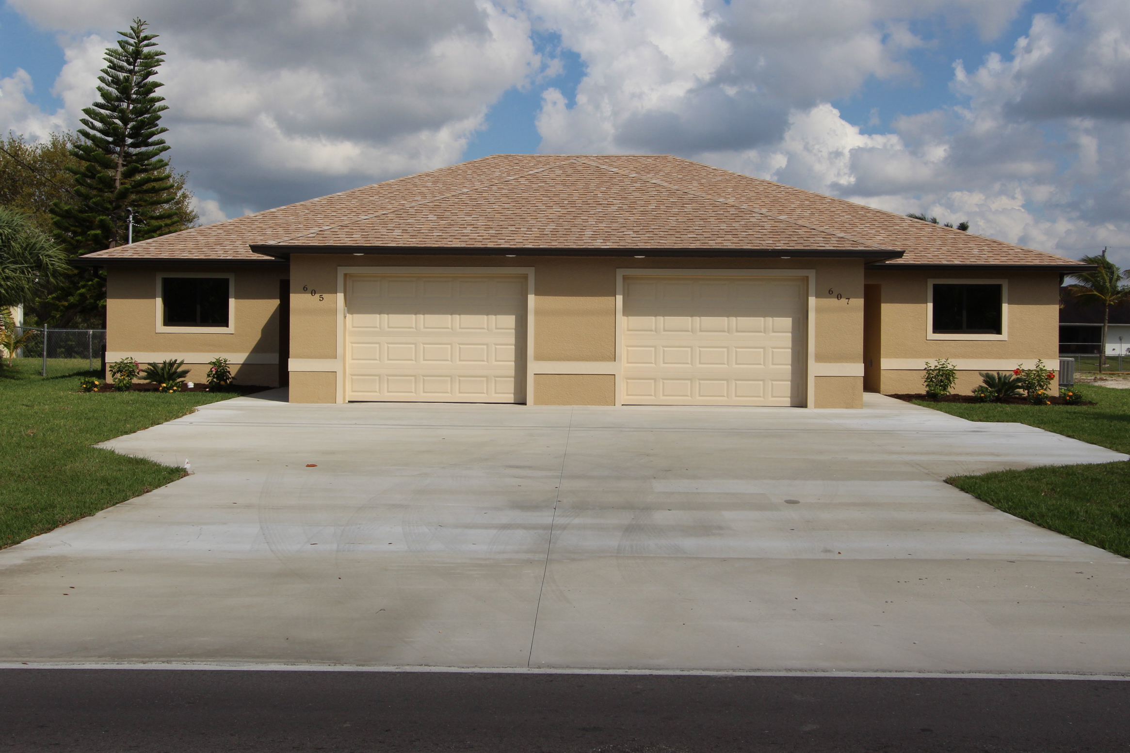 A modern duplex featuring two beige garage doors, tan exterior walls, and a large concrete driveway under a partly cloudy sky—perfect for those seeking homes for rent in Naples.