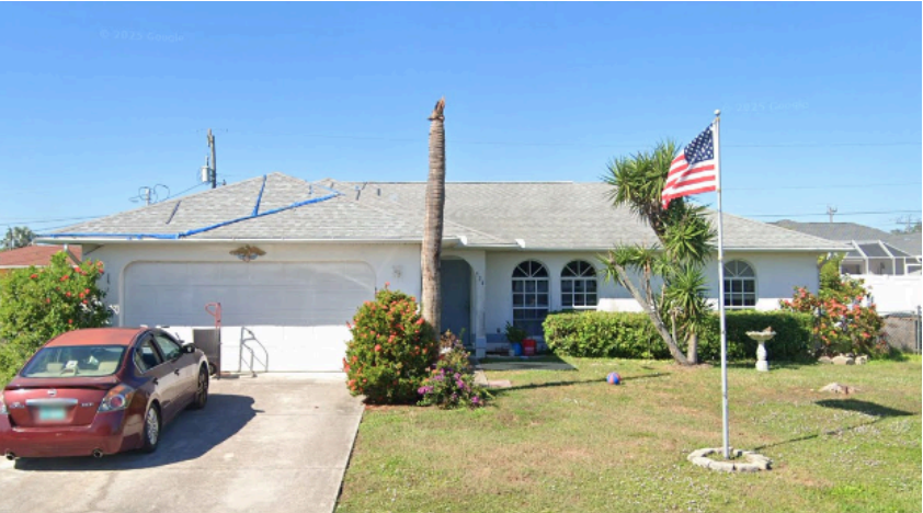 A single-story white Jacaranda house with a gray roof, one bare palm tree, a red car parked in the driveway, and an American flag on a pole in the front yard—featuring 4 bedrooms for comfortable living.