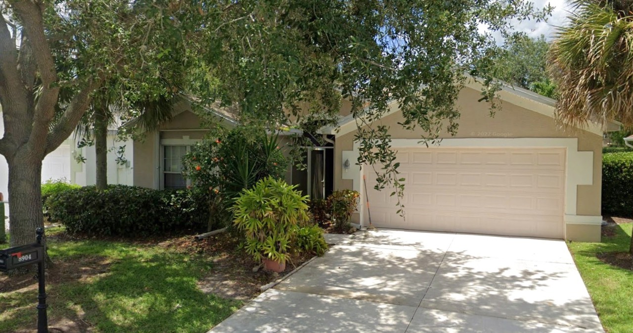 Single-story Fort Myers wholesale property with a light-colored exterior, two-car garage, shaded driveway, front yard with plants and trees, plus a black mailbox at the edge of the lawn.
