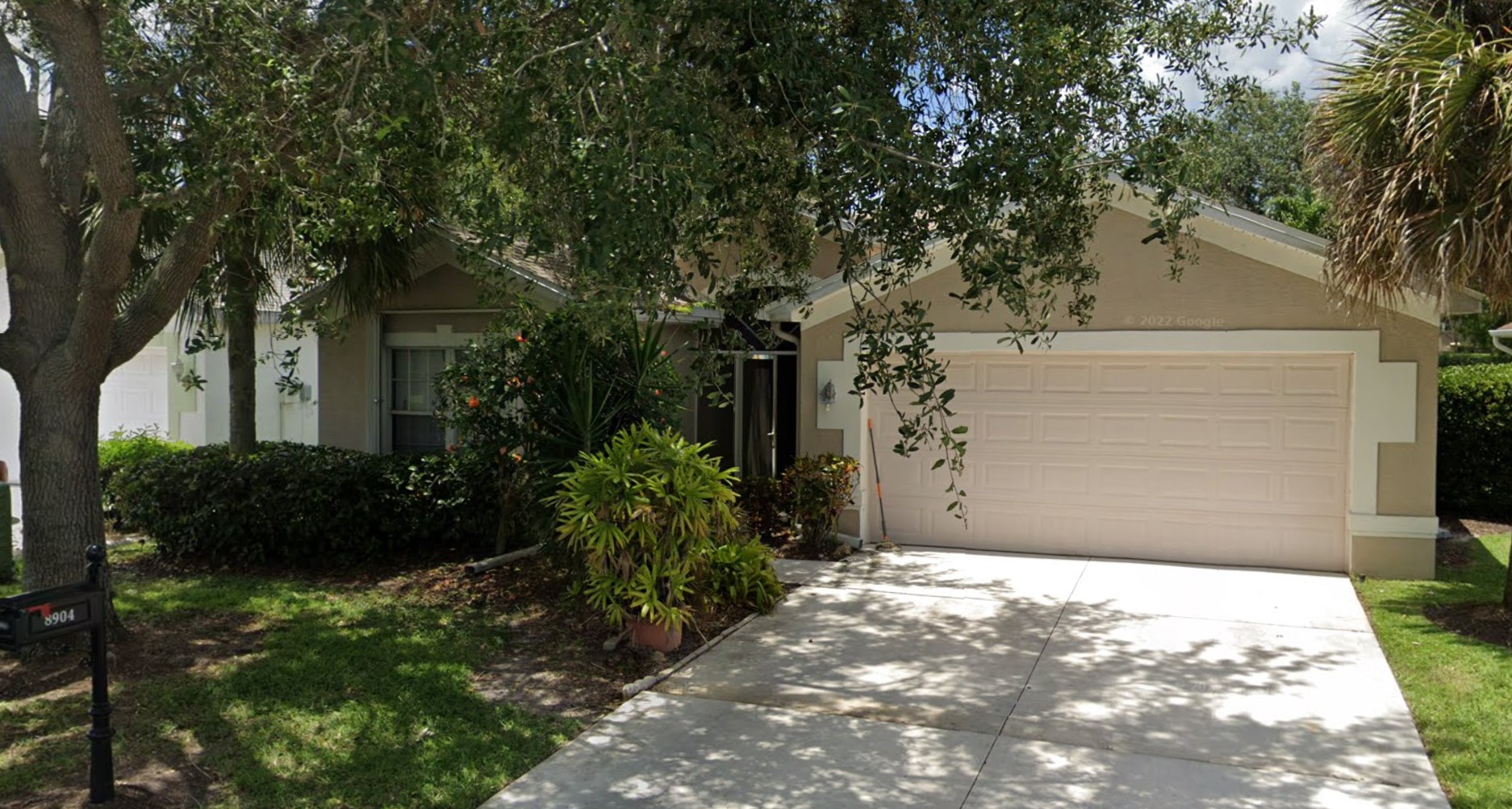 Single-story beige house with a two-car garage, surrounded by trees and greenery, featuring a concrete driveway and a black mailbox at the curb—a prime wholesale opportunity.